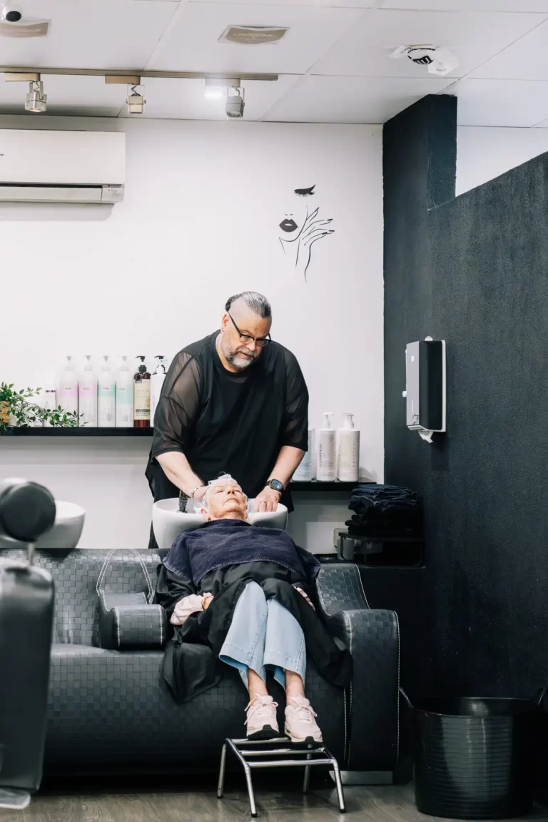Barber washing an older client's hair at a salon station with products on a shelf in the background.