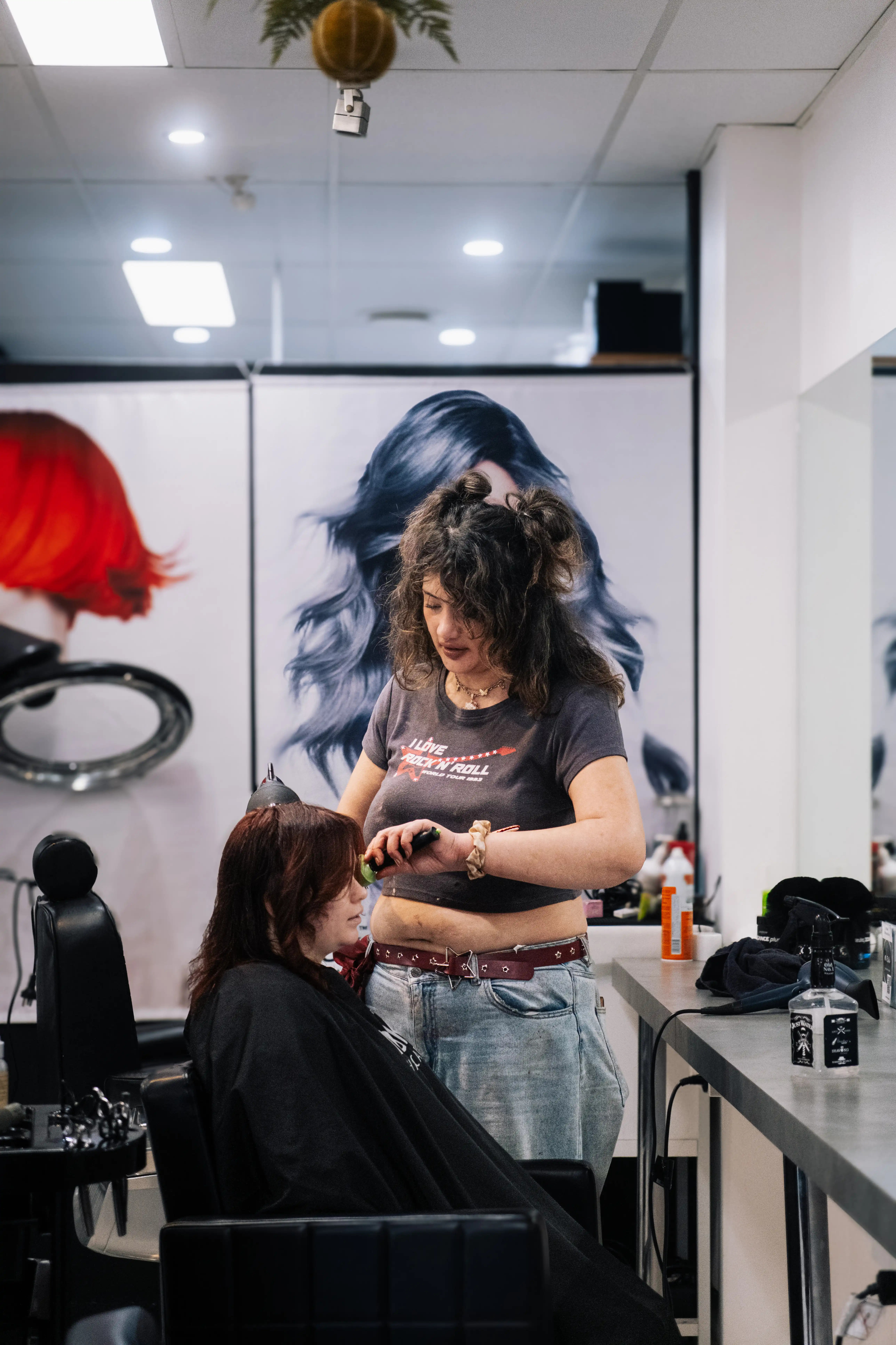 Hairdresser standing and cutting a client's hair in a modern salon, with large poster of blue-haired artwork behind them