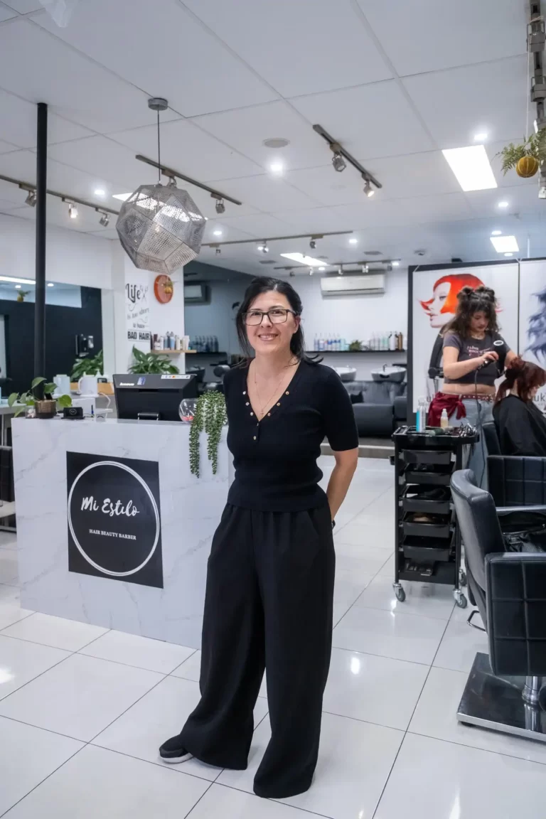 Smiling woman in a black outfit stands in a bright hair salon near a marble reception desk with a 'Mi Estilo' sign. A stylist works in the background.