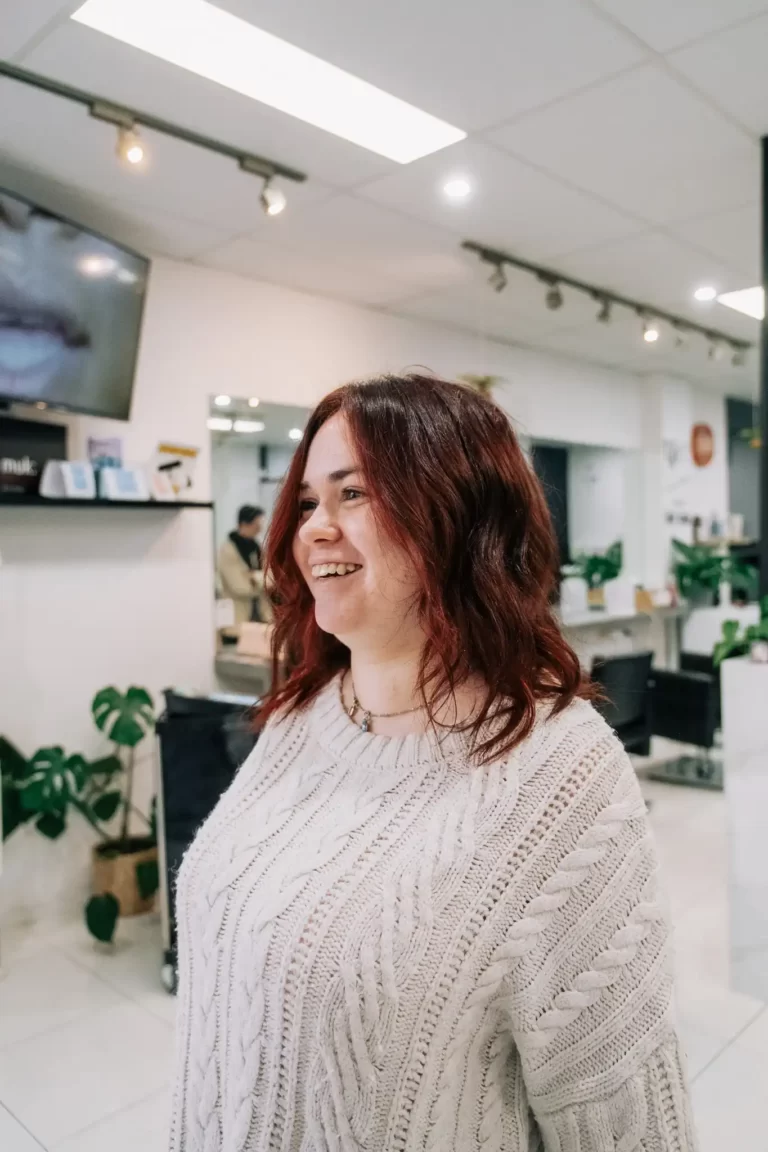 Smiling woman with shoulder-length red hair wearing a white knit sweater in a bright office or shop interior.