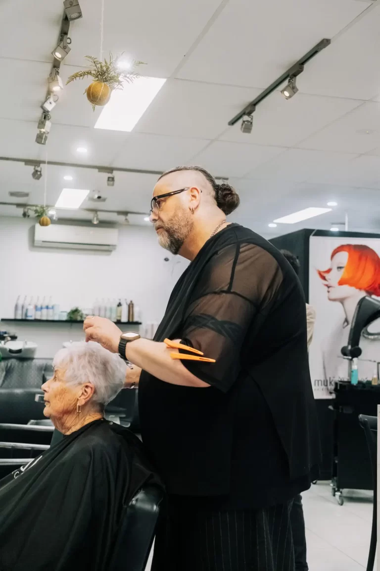 Barber with a bun cutting an elderly woman's hair in a modern salon, holding orange scissors nearby her cape.
