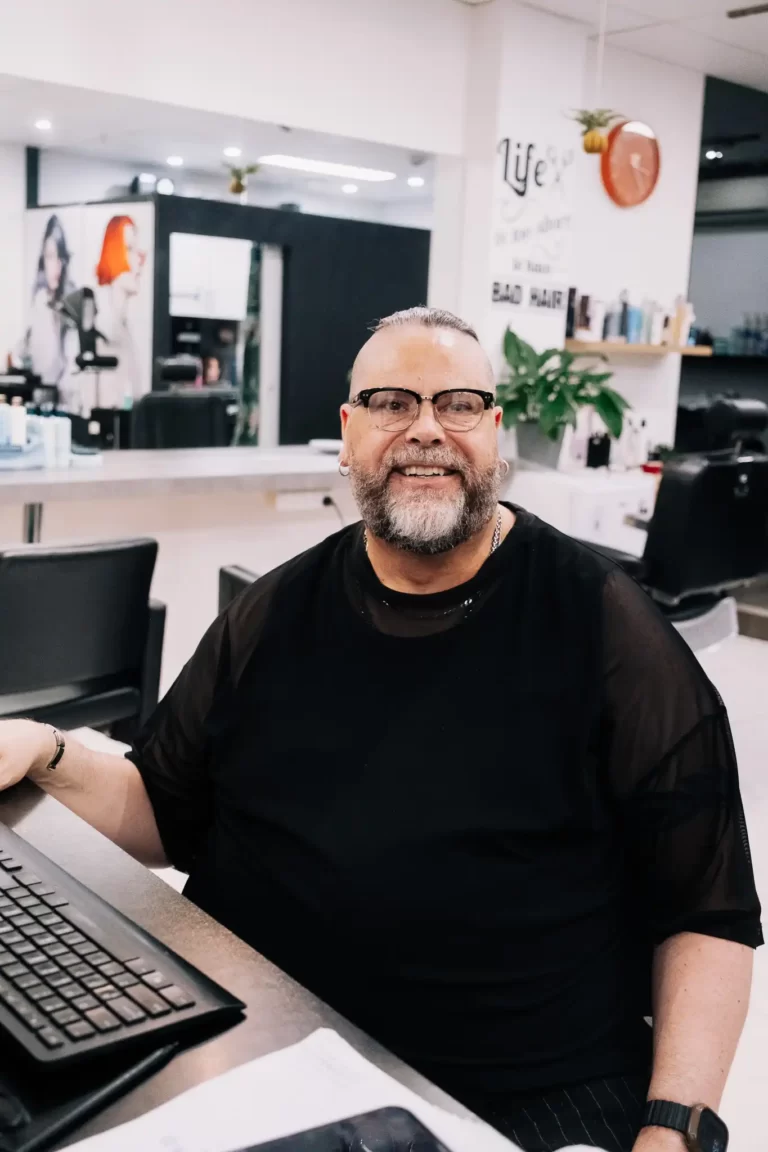 Smiling man with glasses and a gray beard sits at a front desk in a bright salon or office space, a keyboard in view nearby.