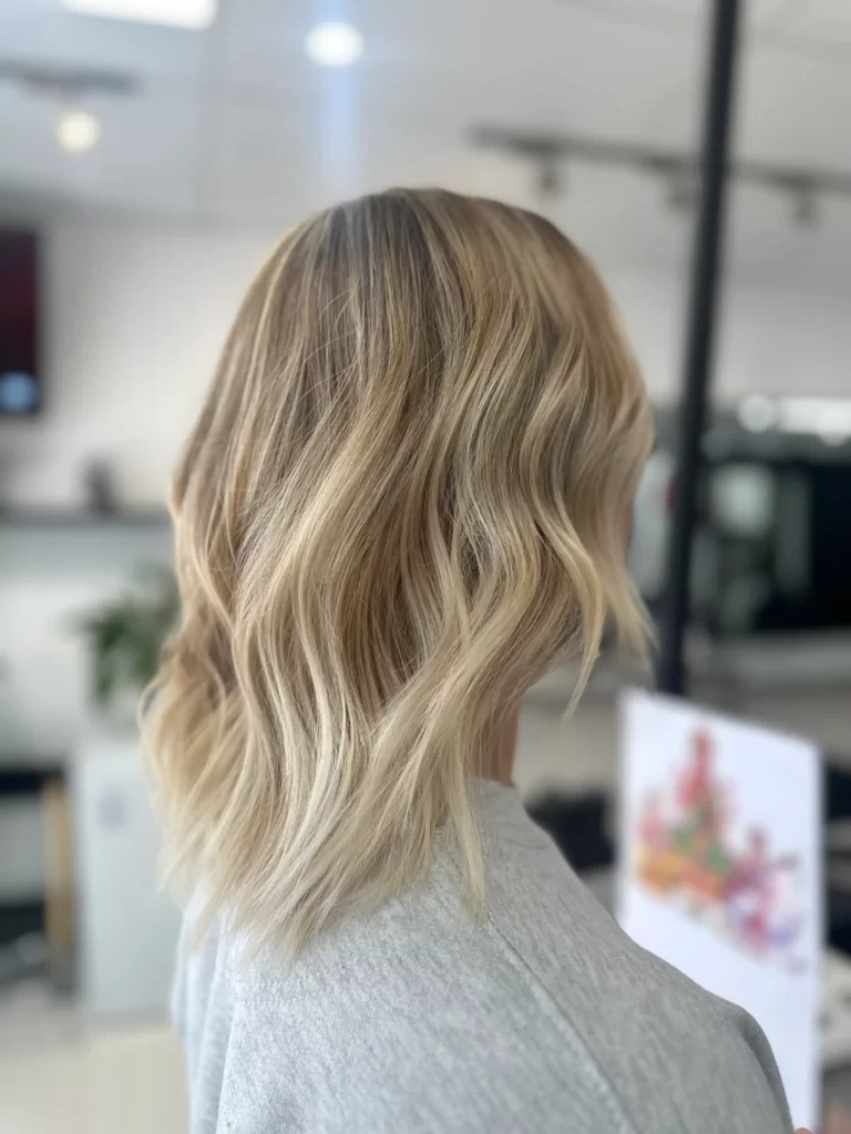 Back view of a person with long, wavy blonde hair wearing a gray sweater in a bright indoor space.